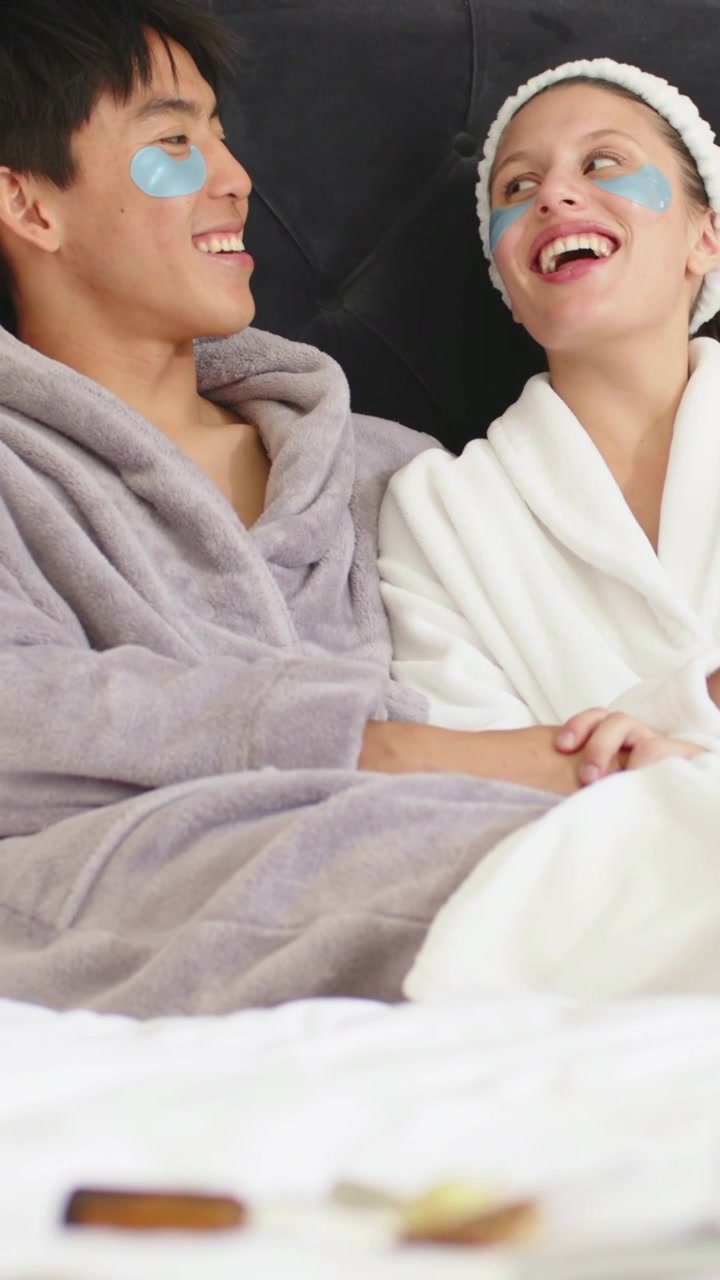 Vertical video: Smiling couple in robes on bed, partner entering left and joining, sharing patches