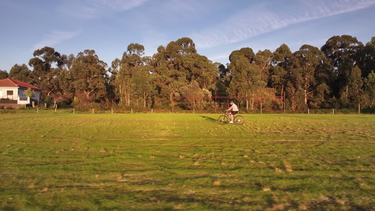 una persona viaja en bicicleta a través de un vasto campo cubierto de hierba rodeado de árboles, bañado por la cálida luz del sol de la hora dorada. un paisaje rural pacífico.