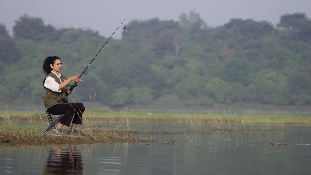 Seorang wanita Asia Selatan memancing di tepi danau yang tenang, duduk di kursi dan memegang pancing, menikmati alam pedesaan yang damai di luar ruangan dalam pemandangan siang hari.