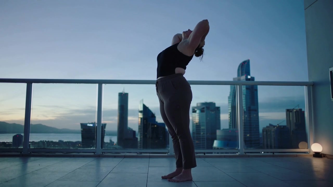 Donna che si inarca all'indietro con top nero e pantaloni grigi, che si riscalda all'alba su un balcone con vista sullo skyline e una lampada accesa.