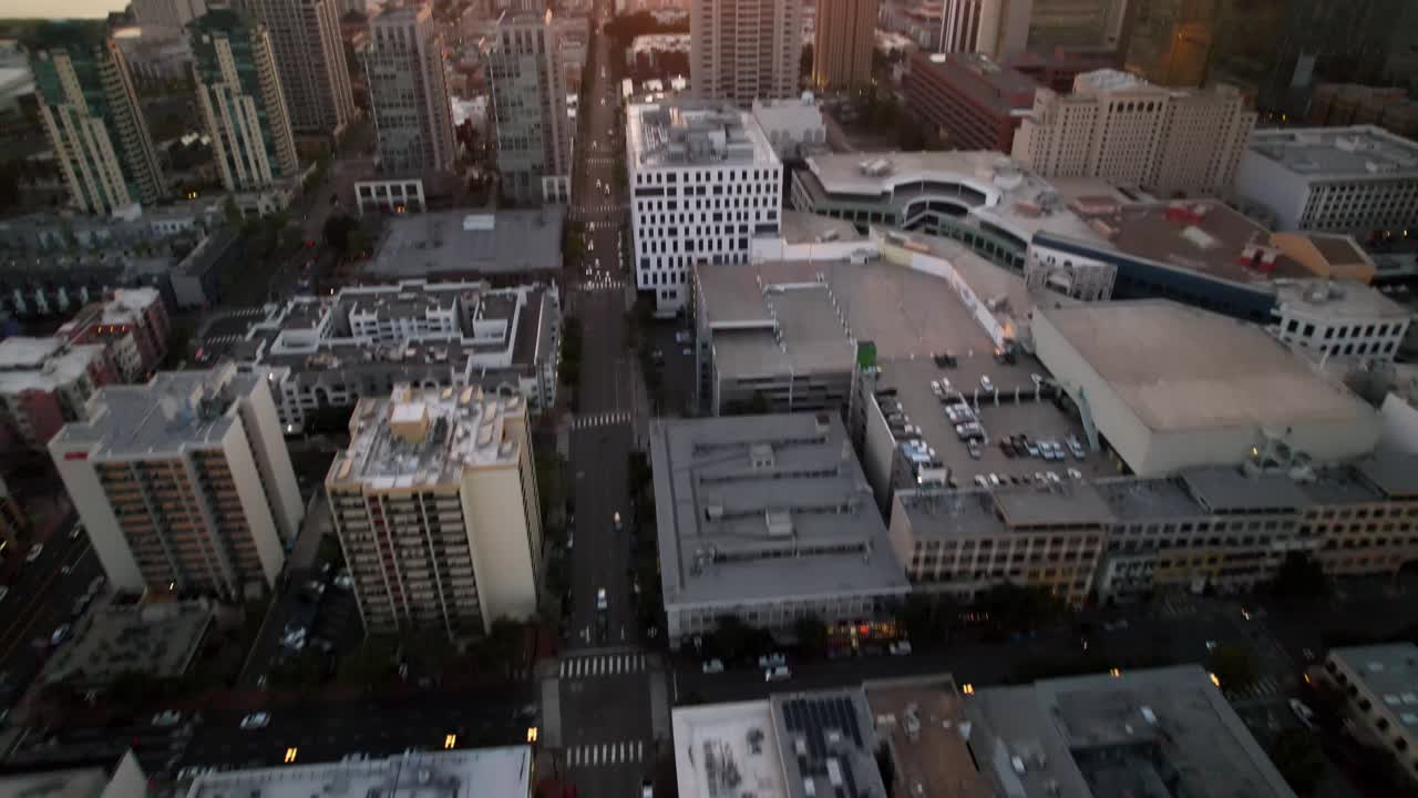 Tomada alternativa de um drone sobrevoando os edifícios do centro de San Diego, com a câmera inclinando para cima para revelar o porto e o pôr do sol.
