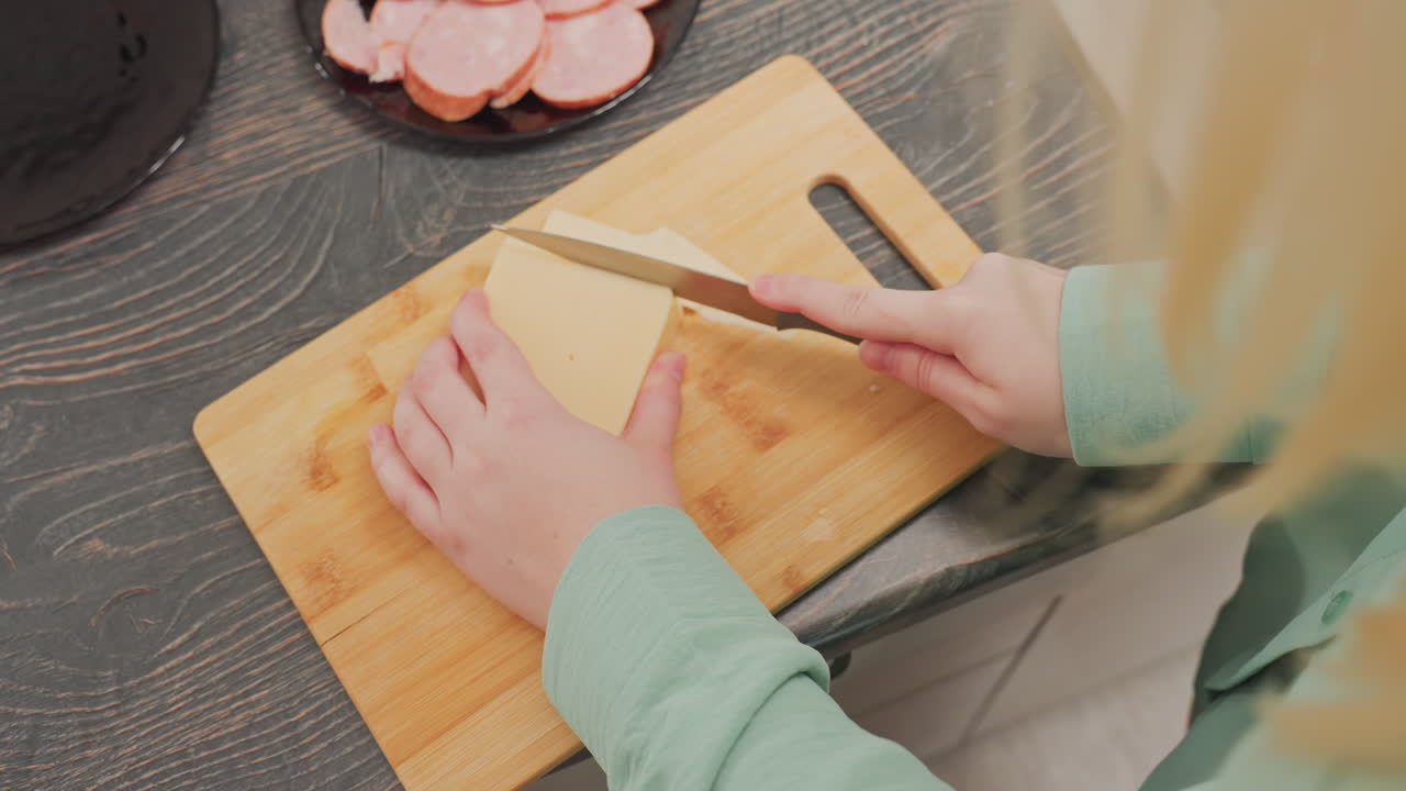 overhead view of woman in green long sleeve slicing block butter with knife on wooden cutting board in kitchen, with sliced sausage arranged on black plate in background on dark wood countertop