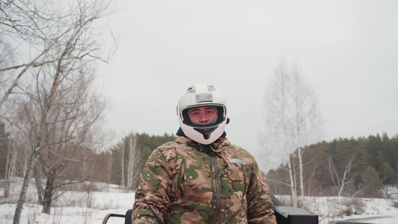 Pilota caucasico esplora un sentiero innevato su un ATV rosso, indossa una giacca mimetica e un casco bianco, ispeziona il terreno e l'orizzonte, preparandosi per un percorso estremo. Postura all'erta e radio portatile visibile.