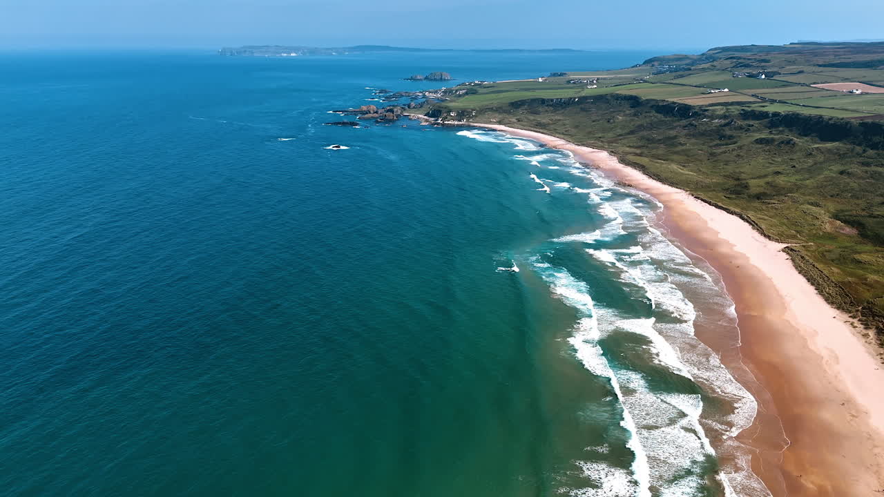 White waves rolling on the sandy beach at the coast of Ireland. Sunny view of a beautiful waterscape and green shore from top view.