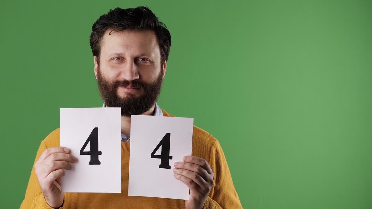 Man holding number 44 sign. Cheerful white bearded good-looking man in yellow sweater shows number forty-four on white sheet. Chrome key, green screen, copy space