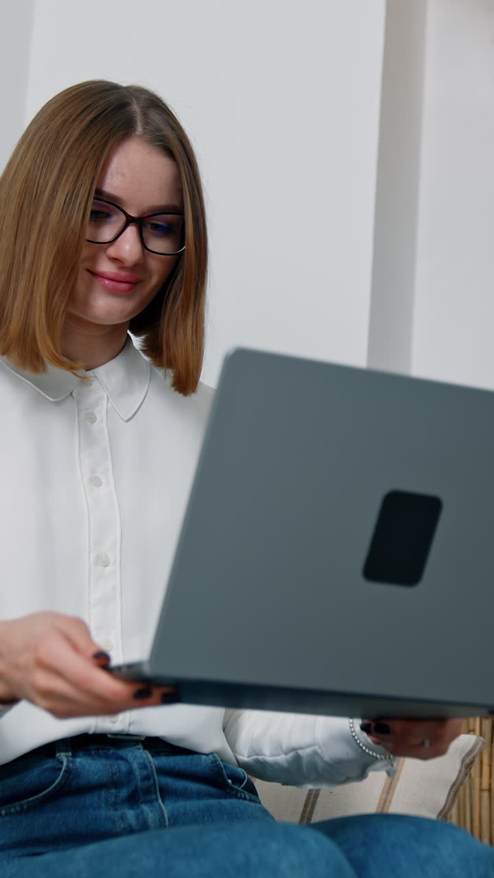 Comfortable workplace at home. Brunette short-haired woman wearing glasses sits working on her laptop. Low angle view. Vertical video