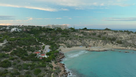 a serene aerial view of konnos beach in cyprus, featuring a secluded sandy cove with crystal-clear turquoise waters, surrounded by lush greenery and rugged cliffs, large hotel overlooking the bay