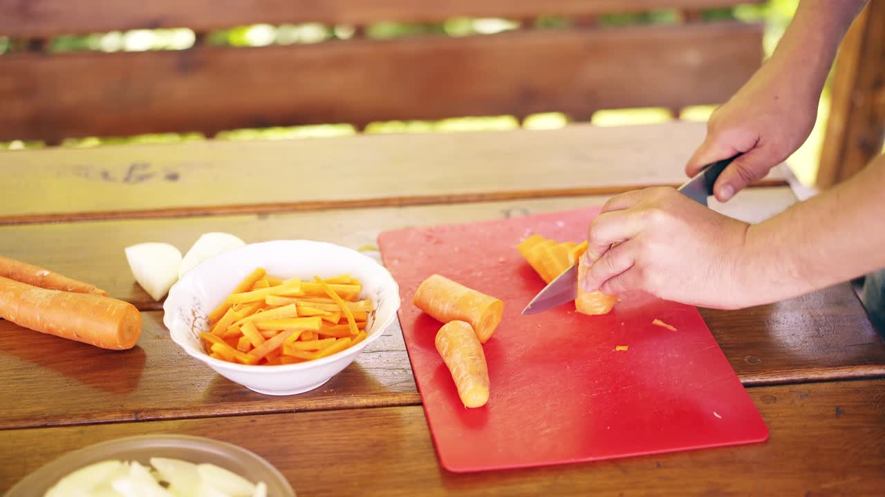 el tipo de primer plano está cocinando pilaf es en una casa de verano corta zanahorias frescas en una tabla de cortar roja