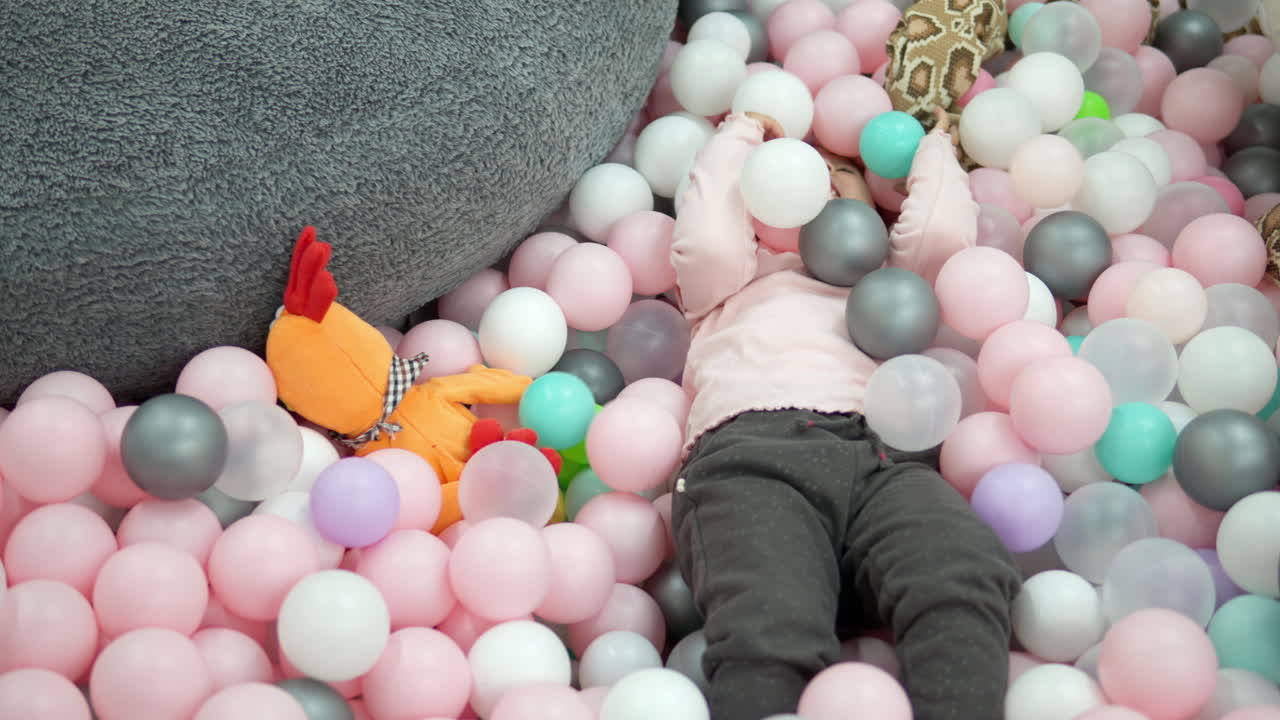 명랑한 어린 소녀가 흰색, 분홍색, 회색 플라스틱 공에 누워 있는 마른 수영장에서 놀고 있습니다.
