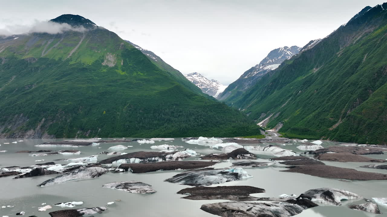 Amazing verdant mountains with a river with melting ice. Snow-capped rocks at backdrop. Alaska wild nature
