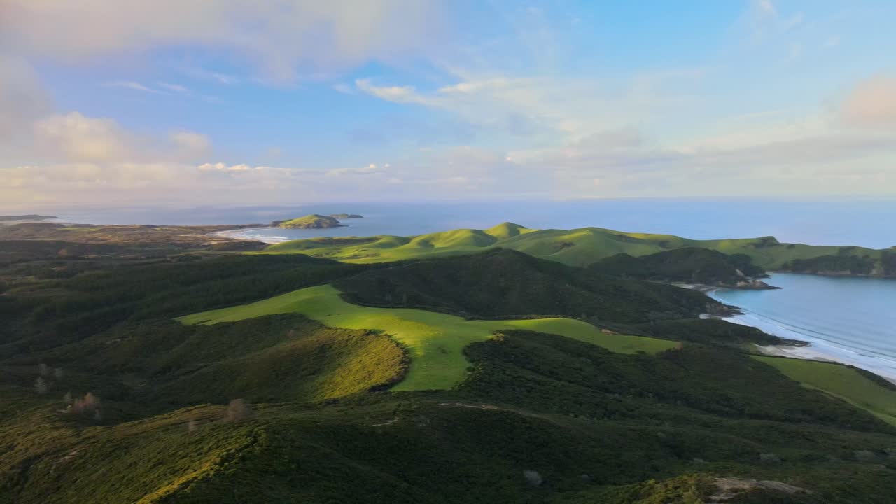 Sideways drone shot at golden hour over Pukenui coastline. Sunny and cloudy sky with ocean views, rugged shoreline, and lush green grasslands glowing in warm evening light