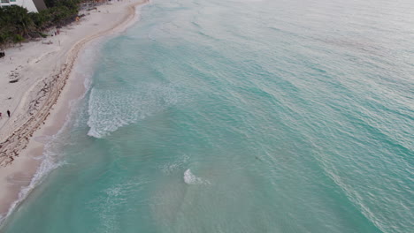 Toma-Aérea-De-Agua-Azul-Cristalina-Con-Olas-Rodando-En-Una-Playa-De-Arena-Blanca-En-Cancún,-México