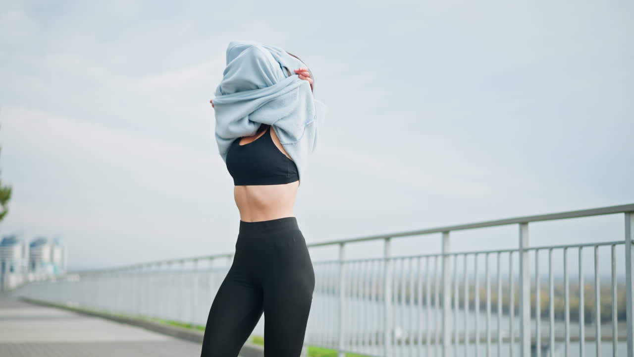Woman standing in front of iron fence, removing her sweater under sunny weather, ideal for fitness, outdoor, and lifestyle moments in a bright, active environment