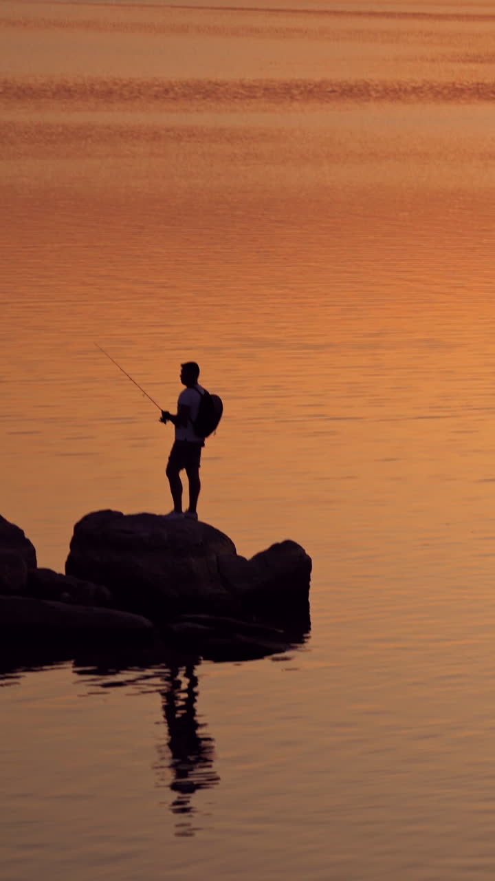 Silhouette of a fisherman with rod on the orange water background. Man with fishing rod is standing on a rock at a beautiful sunset in water. Vertical video