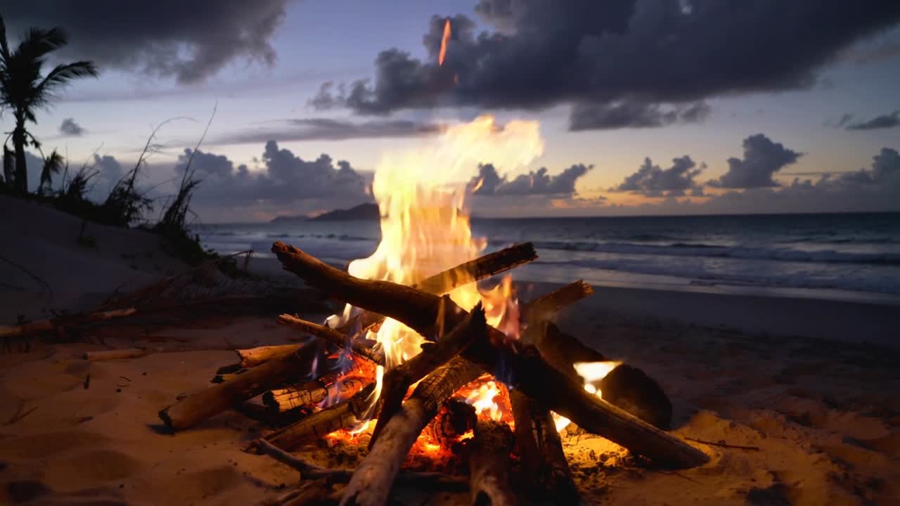 Fogueira queimando em praia arenosa ao pôr do sol, brisa do mar fazendo as chamas tremularem, brasas subindo, viagem.