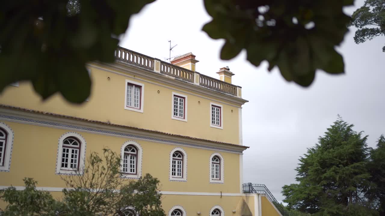 Dolly sideways shot of antique palace building during a cloudy day, tree leaves in the foreground