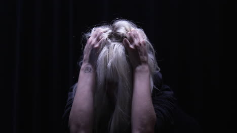 blond depressed woman holding her head, scratching, pulling hair desperately and nervously in a dimly-lit room illuminated by a single overhead light