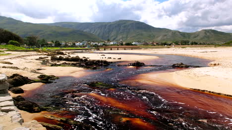 Brown-tannin-rich-Kleinmond-lagoon-water-flows-over-beach-toward-ocean,-scenic