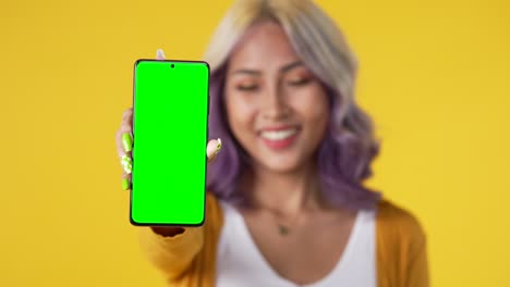 joven asiática feliz mostrando un teléfono inteligente con pantalla verde aislada sobre un fondo amarillo