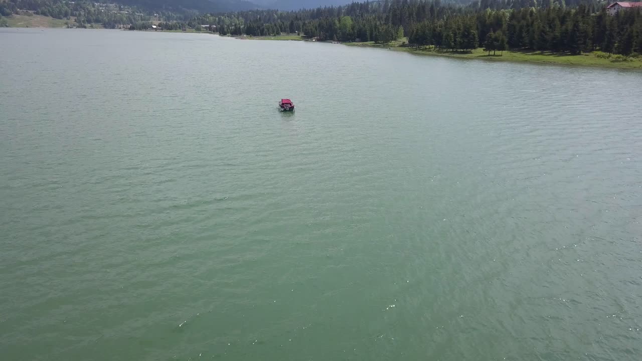 Panoramica aerea di un lago con una barca che galleggia al centro e montagne sullo sfondo.