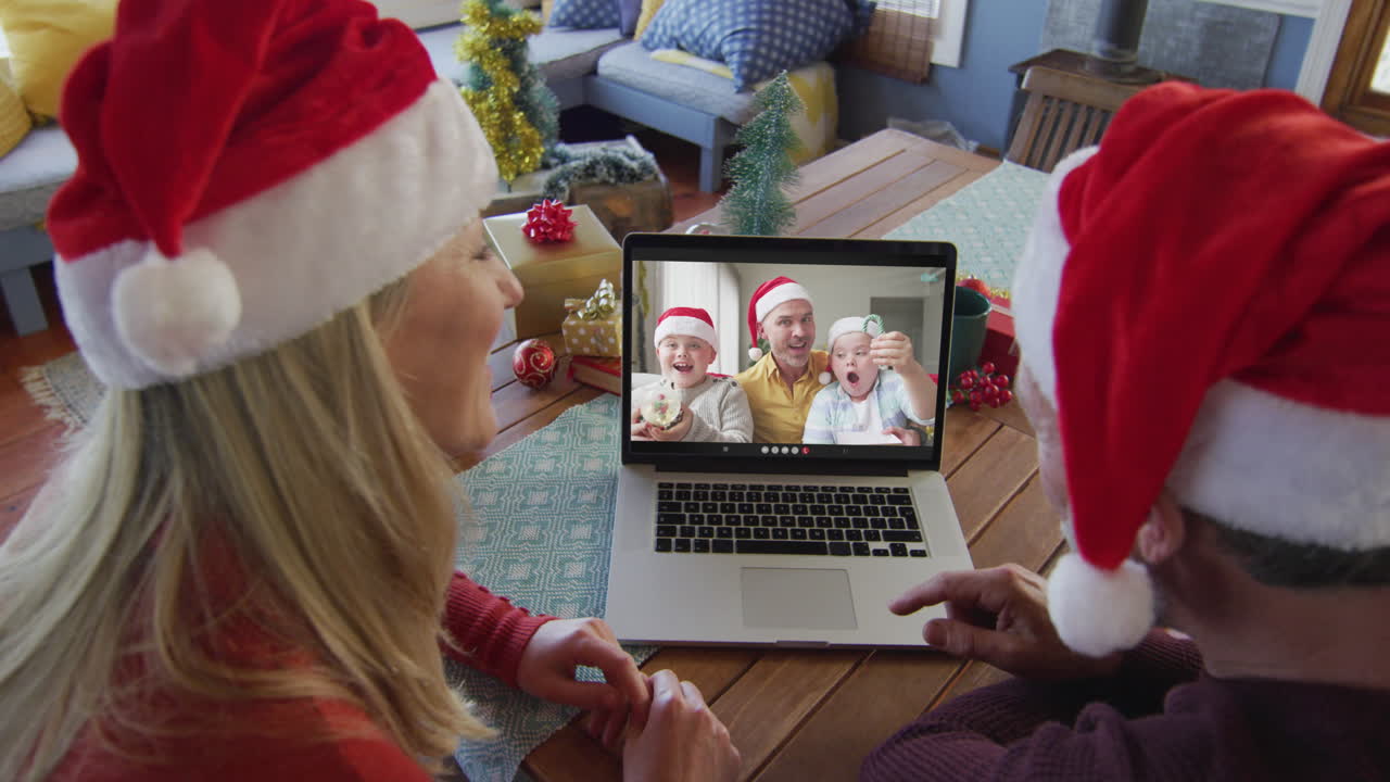 pareja caucásica sonriente con sombreros de santa usando una computadora portátil para una videollamada de navidad con la familia en la pantalla