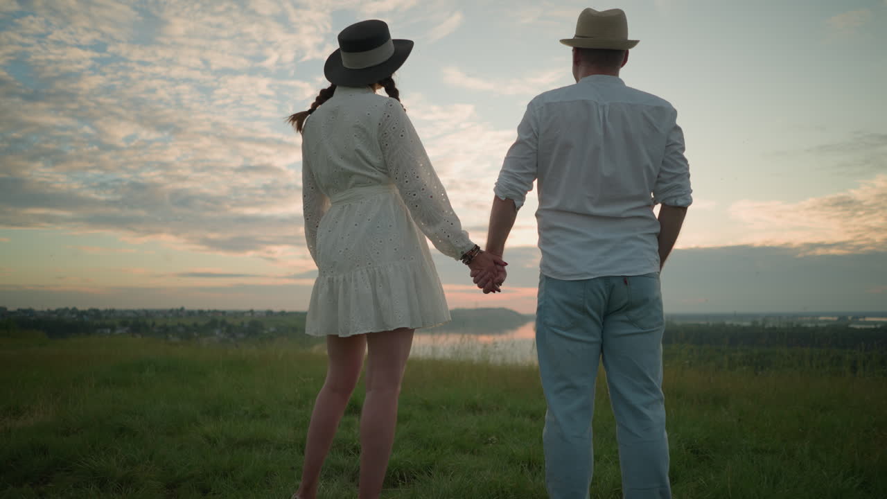 Newlyweds stand hand in hand on a grassy hill at sunset, gazing at the sky. The man, dressed in a white shirt, hat, and jeans, holds hands with the woman in a white dress and hat