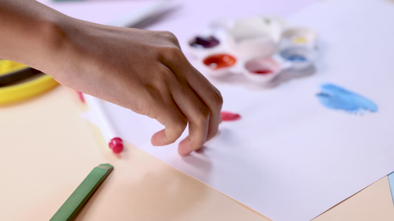 Hand applies red paint fingerprint on white paper, surrounded by palette, pencils, and soft lighting