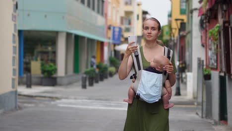 with her infant in a kangaroo backpack, a young woman takes photos on a mobile phone during her journey. she strolls along, occasionally looking at the phone screen