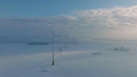 aerial view of wind turbines generating renewable energy in the wind farm, snow filled countryside landscape with fog, sunny winter evening with golden hour light, wide drone shot