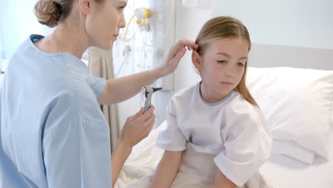 caucasian female doctor using otoscope to check ear of girl patient in hospital bed, slow motion