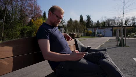 caucasian man uses mobile phone, sits on a bench outside on sunny day