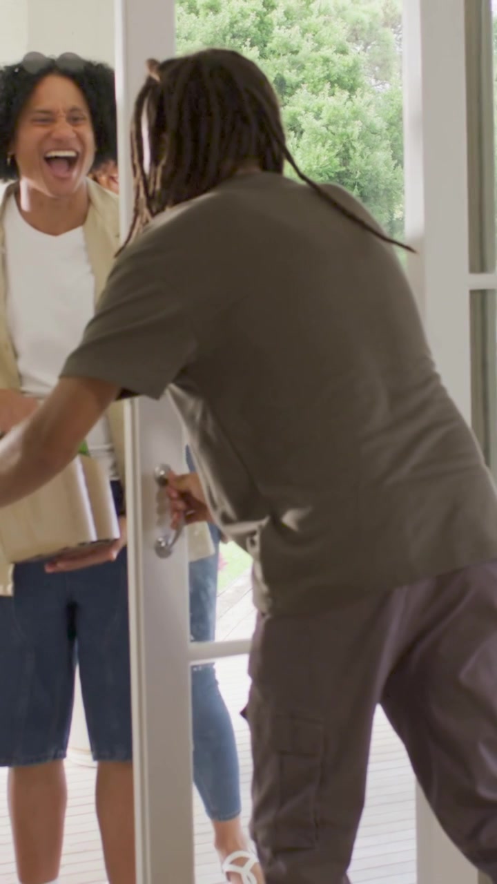 Vertical video: Opening glass door host with dreads greeting guests arriving with carrier on patio
