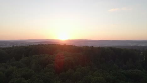 Material-De-Archivo-De-Vuelo-Rápido-Sobre-Un-Bosque-Enorme-En-Una-Puesta-De-Sol-Naranja-Y-Dando-Hermosos-Destellos-Solares-En-Las-Imágenes-Con-Montañas-En-El-Fondo