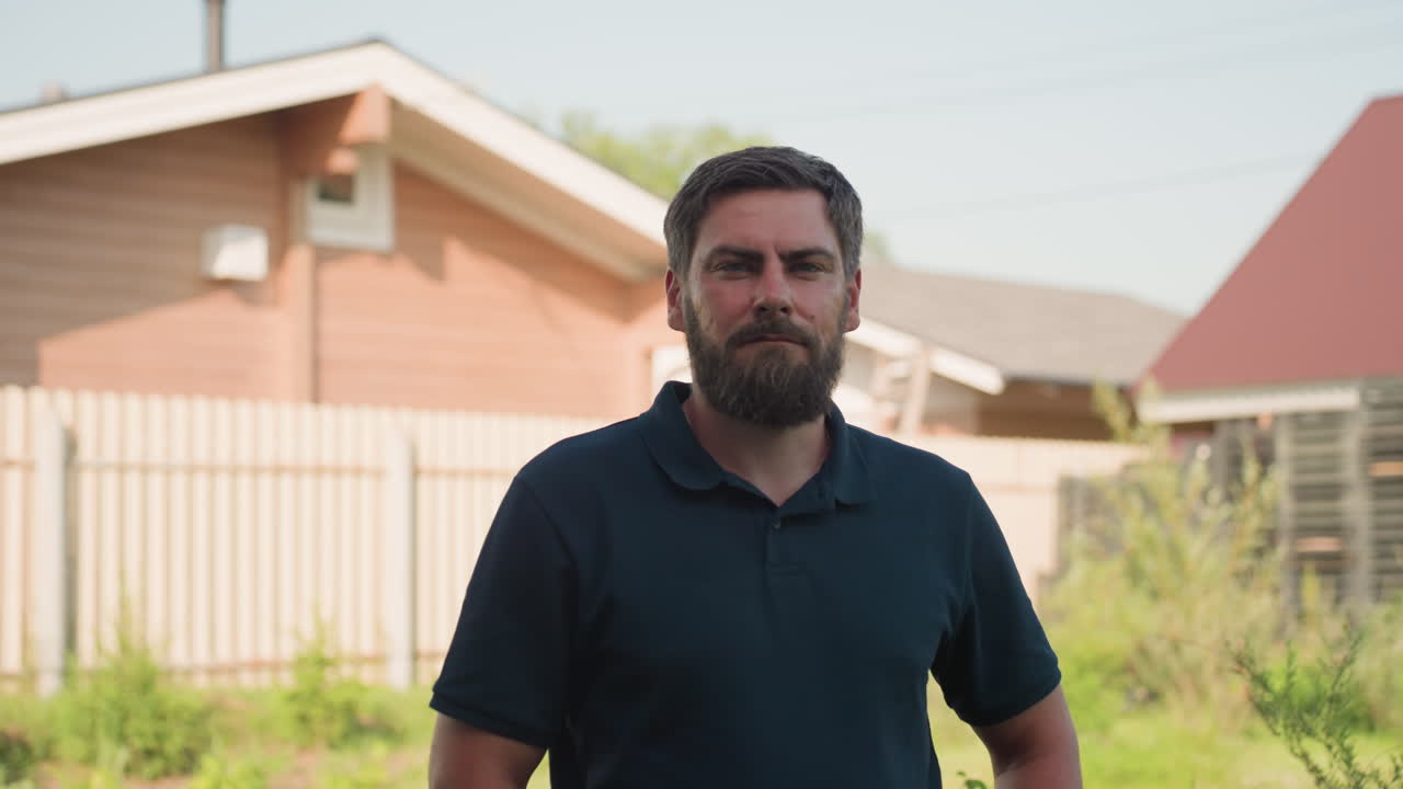 Bearded farmer in dark polo stands with hands on waist then removes hand to hold trowel from belt pocket, standing beside blue drum with wooden crate in sunny backyard