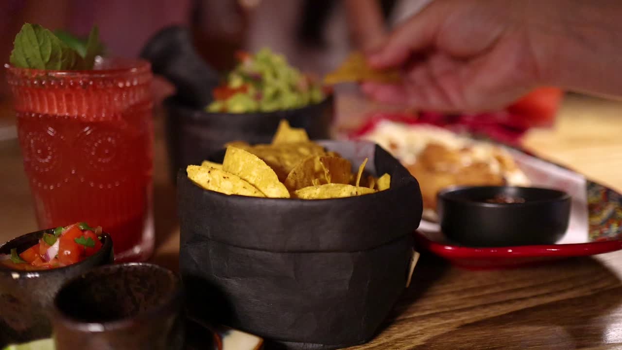 Una mano afferra patatine nacho da un cestino, intingendole nel guacamole e nella salsa, con vivaci piatti messicani e un cocktail rosso su un tavolo di ristorante illuminato calorosamente.