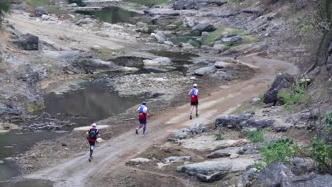 Group-of-marathon-athletes-running-a-charity-race-on-dirt-road-along-river-creek-through-forest-jungle-in-rural-beautiful-nature-of-Ecuador-50-fps