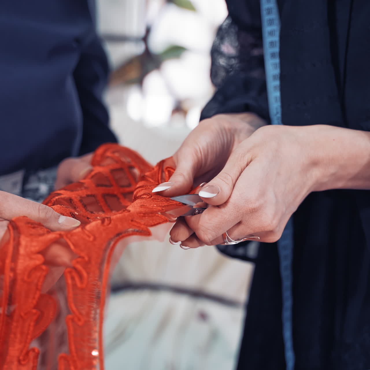 Professional dressmaker is cutting red lace with scissors with the help of a tailor in atelier. Woman's hands cutting out sequin and beaded red lace along its edge following the contour.