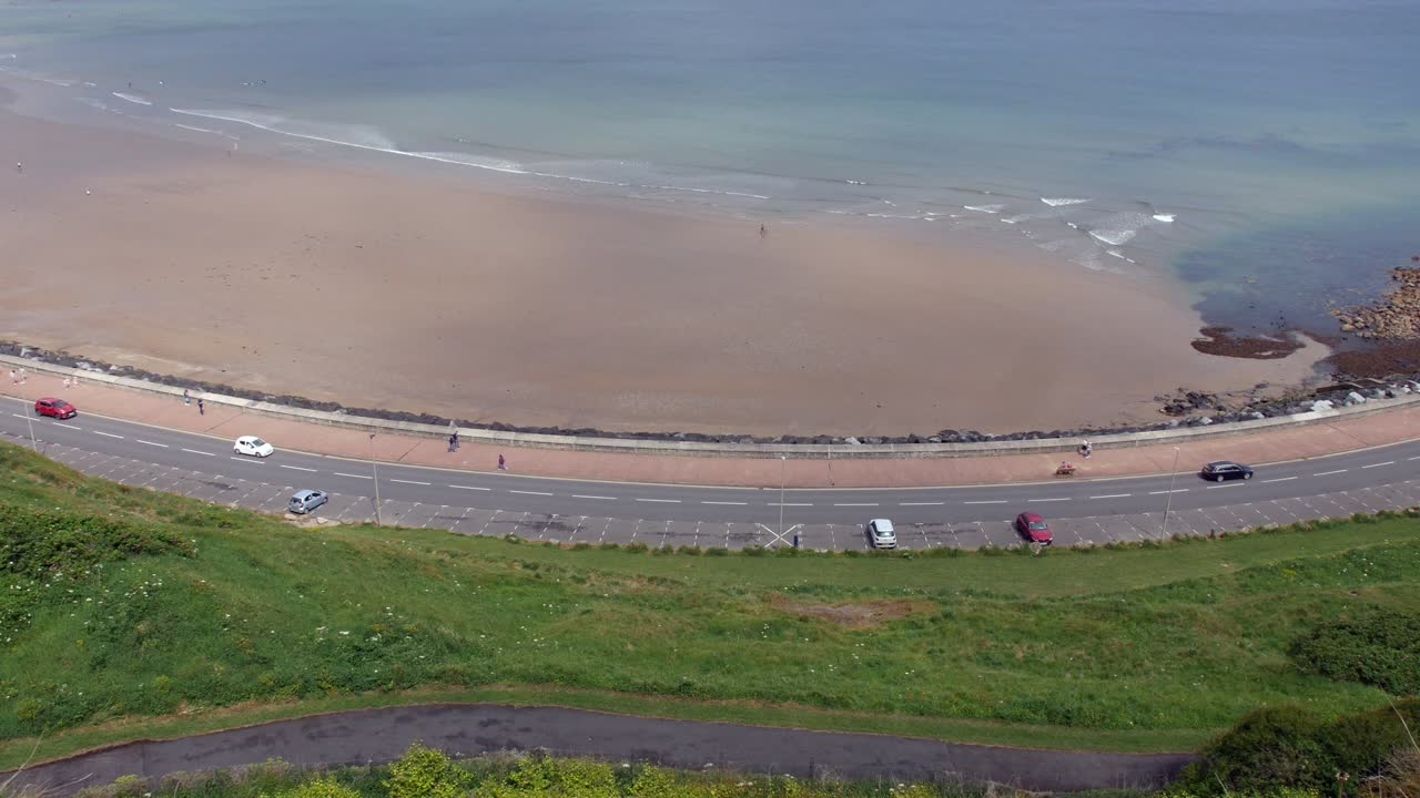 A beach stretches along the shoreline with gentle waves lapping against wet sand, while cars are driving on a coastal road curving below grassy cliffs. Pedestrians are walking on the seaside promenade