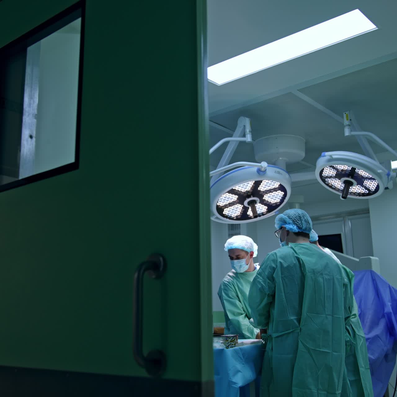 Puerta corredera entreabierta a un quirófano moderno con una operación en curso. Tres cirujanos trabajando en la mesa de operaciones.