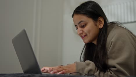 Nahaufnahme-Eines-Glücklichen-Brünetten-Mädchens-In-Einem-Braunen-Pullover,-Das-Auf-Ihrem-Grauen-Laptop-Tippt,-Während-Es-Tagsüber-In-Einem-Modernen-Apartment-Im-Schlafzimmer-Auf-Dem-Bett-Liegt