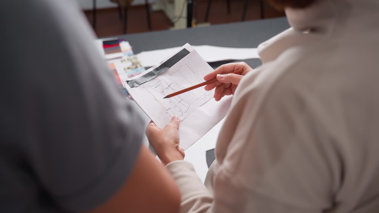 Back view of coat design sketch held by creative as she uses pencil to make precise markings during clothing development session in modern workspace surrounded by fabric samples and paper sheets