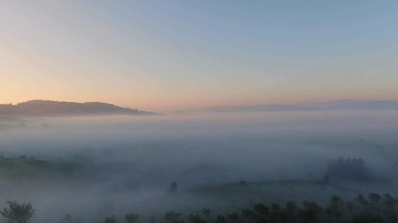 Landscape with fog The sky is blue with some pink tints The fog covers the valley below There are some trees that are above the fog line