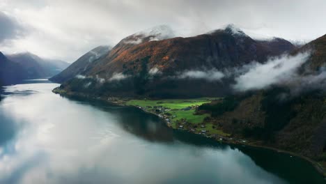 aerial shot of a peaceful village beside a fjord, surrounded by mist-covered mountains and calm waters