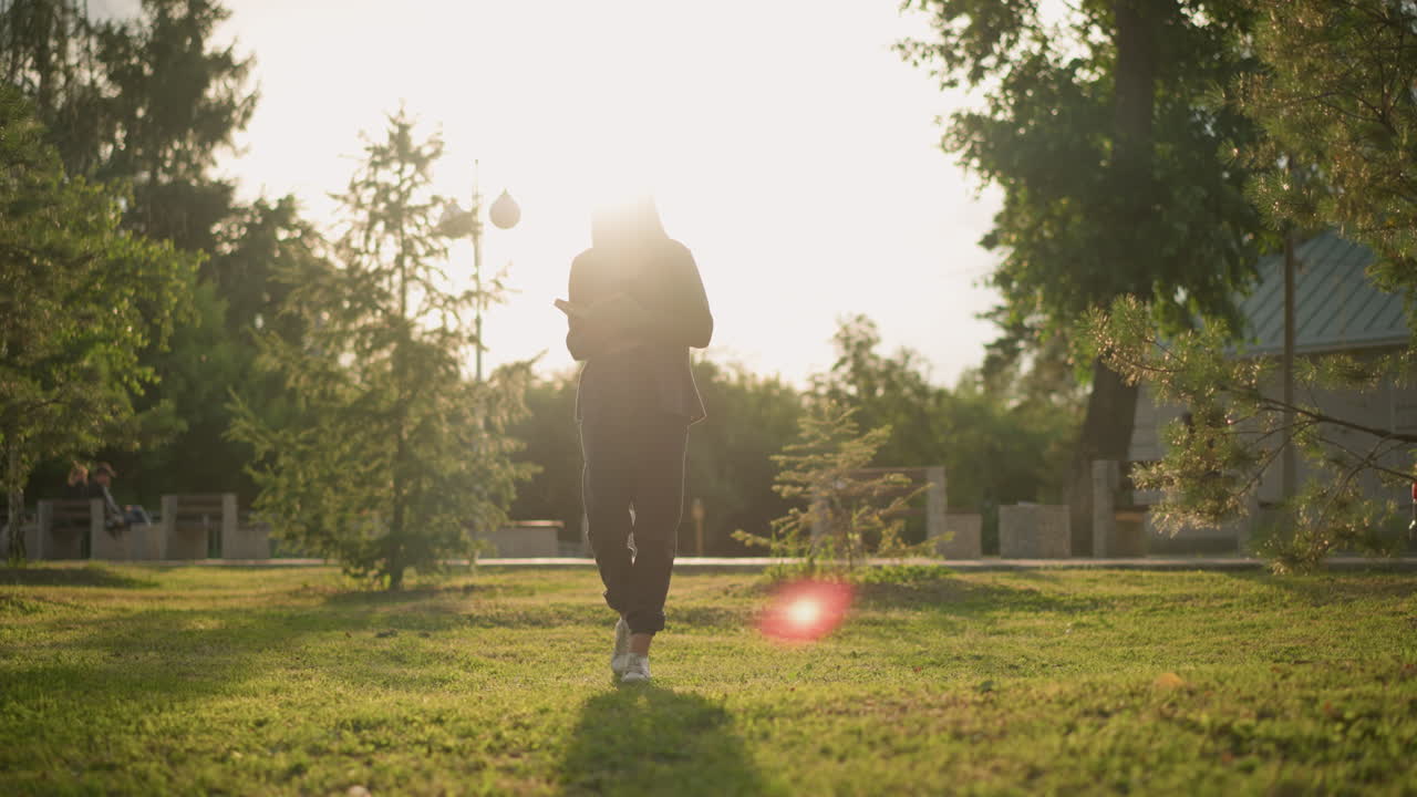 회색 옷을 입은 여인이 잔디에서 걸어가며 책을 읽으며 빛이 그녀를 둘러싸고 밝은 반임으로, 나무와 건물과 함께 배경에 차의 부분적인 시야를 만니다.