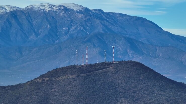 Panoramica teleobiettivo su torri di comunicazione su una collina arbustiva, sovrastate da un'imponente parete montuosa andina innevata che riempie l'intero sfondo