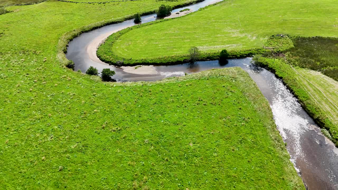Drone camera glides above a winding river through lush green farmland in Glen Clova, Scotland, under bright daylight with smooth, steady motion