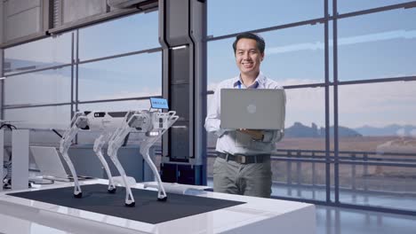 cuerpo lleno de un trabajador profesional masculino asiático de pie con su portátil en una fábrica de alta tecnología, perro robot, está mirando a la cámara con una sonrisa