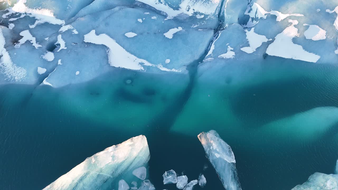 Drone top down shot of large floating icebergs drifting in cold blue glacial water in Iceland. Aerial view reveals deep cracks, textures, and shades of ice surrounded by small chunks and shadows.