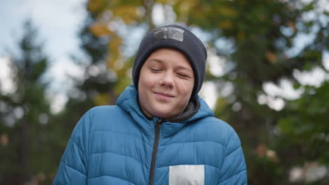 kid in black beanie and blue jacket standing outdoors, smiling and enjoying the moment with a happy expression, blurred greenery and autumn scenery in the background