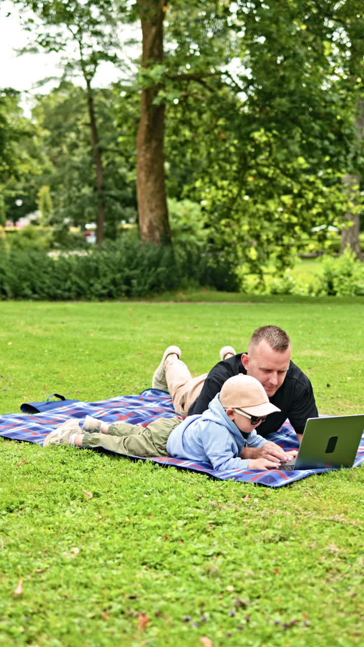 Sødt barn og far tilbringer tid sammen. Lille dreng og hans far kigger på en bærbar computer, mens de sidder på græsset. Far og søn-tid i parken. Glad faderskab og sjov barndom.